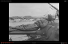 US tank crewman reclining on main gun of Sherman tank LCT 902 in background.png